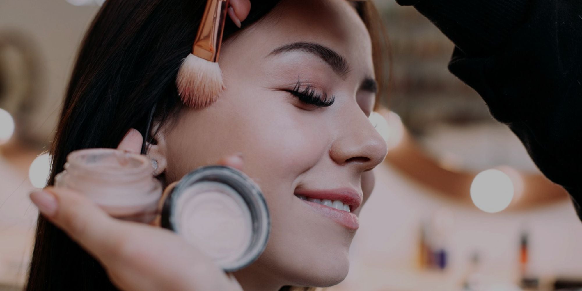 A close-up lifestyle shot of a woman receiving makeup application, with a makeup artist holding a small jar of cream blush and a makeup brush near her temple. The background is a softly blurred, warm-toned indoor setting.